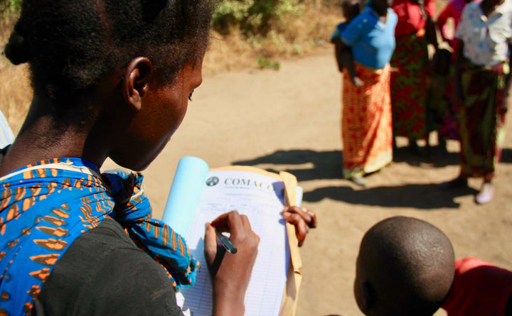 Female African farmer fills in a purchase order form for COMACO. She is near a group of people and next to a child.